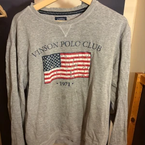 Grå sweatshirt Vinson Polo Club - Säljer en grå sweatshirt från Vinson Polo Club med tryck av amerikanska flaggan och texten 'VINSON POLO CLUB 1971' på bröstet. Tröjan har rund halsringning och långa ärmar. Perfekt för en avslappnad och sportig stil.