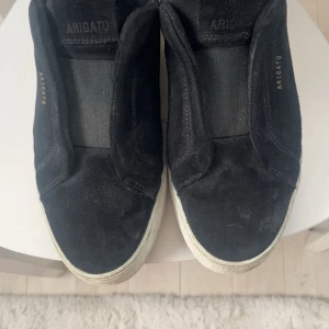 Svarta sneakers från Axel Arigato - Svarta sneakers från Axel Arigato i mocka med vit sula. Skorna har elastiskt band över vristen för enkel påtagning och en stilren, minimalistisk look. Perfekta för dig som gillar klassisk streetstyle med premiumkänsla. Jättefina bra skick! 