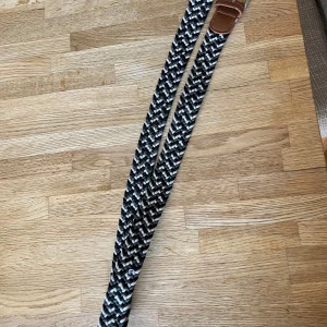 Svartvit flätad bälte med läderdetalj - Säljer ett snyggt flätat bälte i svart, vitt och grått med bruna läderdetaljer vid spännet. Bältet har ett coolt mönster och passar perfekt till jeans för en avslappnad men stilren look.