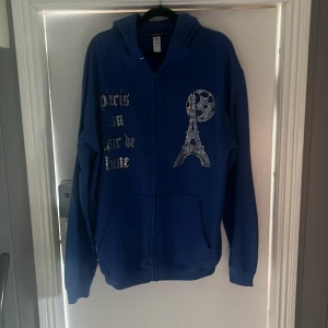 Blå hoodie med strassdetaljer Blinglucid - Blå hoodie från Blinglucid med dragkedja och huva. Framsidan har strassdekor med texten 'paris au clair de lune' och Eiffeltornet i bling. Tillverkad i 80% bomull och 20% polyester. Perfekt för dig som gillar statement-plagg och coola detaljer. Aldrig använd då jag fick L istället för S
