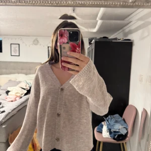 Beige stickad oversize kofta med knappar - Mysig beige stickad oversize kofta med v-ringning och stora knappar framtill. Koftan har en loose passform och långa ärmar, perfekt för lager-på-lager. Enkel och stilren design som funkar till allt.