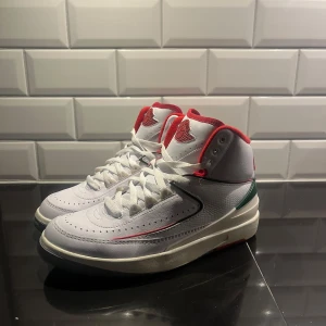 Nike Air Jordan 2 vita och röda - Nike Air Jordan 2 sneakers i vitt skinn med röda detaljer och grönt parti vid hälen. Klassisk hög modell med snörning, rund tå och vadderad plös med Air Jordan-logga. Sulan är vit med rött och grått mönster undertill. Perfekta för dig som gillar retrostil och streetwear.