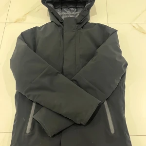 Svart dunjacka från Jack & Jones S - Säljer en svart dunjacka från Jack & Jones i storlek S. Jackan har huva, dragkedja och två fickor med dragkedja framtill. Perfekt för kalla dagar med sin vadderade insida och stilrena look.