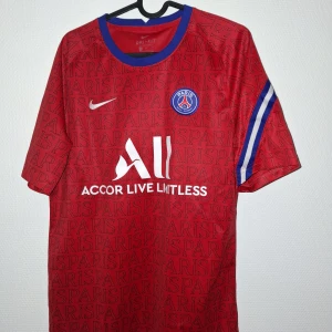 Röd PSG fotbollströja Nike M - Snygg röd PSG fotbollströja från Nike med Dri-Fit material. Tröjan har blå krage och blåvita ränder på ärmarna. All-over print med 'PARIS' och PSG-logga på bröstet. Perfekt för dig som vill sticka ut på planen eller på läktaren.