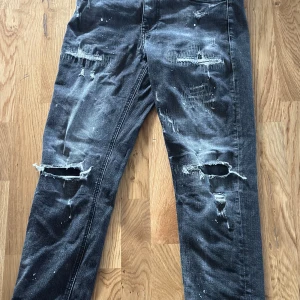 Grå slitna jeans Supply & Demand - Grå jeans från Supply & Demand med coola slitningar och hål på knäna. Jeansen har en tvättad look med ljusa partier och färgstänk, klassisk femficksmodell och rak passform. Perfekta för dig som gillar streetstyle och edgy vibe.