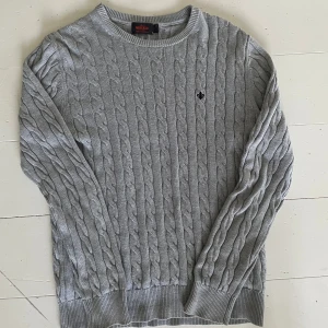 Grå kabelstickad tröja från Morris - En nästan helt oavänd snygg grå kabelstickad tröja från Morris i 100% pima bomull. Klassisk modell med rund halsringning, ribbade muddar och diskret svart brodyr på bröstet. Perfekt nu till hösten🍁🍂 
