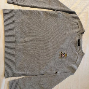 Grå stickad tröja Morris Gents S - Snygg grå stickad tröja från Morris Gents i storlek S. Tröjan har broderad logga på bröstet med detaljer i svart, rött, vitt och guld. Klassisk rund halsringning och ribbade muddar vid ärmslut och nederkant. Perfekt för en stilren look.   Tveka inte att fråga om ni vill ha fler bilder eller undrar något, Mvh Olle