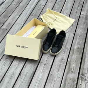 Svarta sneakers från Axel Arigato - Stilrena svarta sneakers från Axel Arigato i skinn med vit platt sula. Låg modell utan snörning, med elastiska paneler för enkel påtagning. Diskret guldfärgad logga på sidan och klassisk rund tå. Perfekta för en clean och modern look. Har dock blivit lite lagade där bak annars är det perfekta.