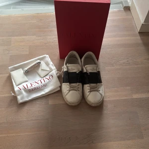 Valentino Garavani Open sneakers vit/svart - Snygga Valentino Garavani Open sneakers i vitt skinn med bred svart rem över vristen. Klassisk rund tå, platt sula och diskret logga på plösen. Skorna levereras med originalpåse och röd box. Perfekta för dig som gillar exklusiva och trendiga sneakers.