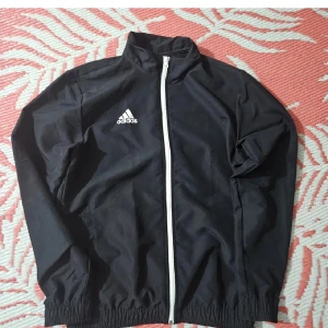 Svart vindjacka från Adidas - Svart vindjacka från Adidas med vit dragkedja och klassisk logga på bröstet. Jackan har hög krage, långa ärmar och resår vid ärmslut och midja. Perfekt för en sportig look och enkel att slänga på sig när det blåser ute.