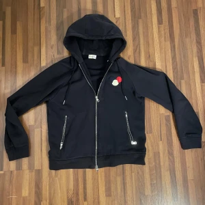 Moncler zip up - Svart hoodie från Moncler med hel dragkedja, huva och två fickor med dragkedja. På bröstet finns Monclers ikoniska brodyr i blått, vitt och rött. Tröjan är i storlek L men den har en ganska annorlunda passform och den passar mig som vanligtvis har M, den passar nog även S.