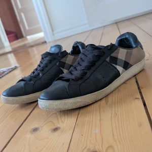 Burberry sneakers med rutigt mönster - Snygga sneakers från Burberry med klassiskt rutigt mönster i beige och svart på sidorna. Skorna har svarta skinnpartier, svarta snören och en vit sula. Använda men i bra skick fortfarande