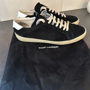 Svarta Saint Laurent sneakers i mocka - Snygga svarta sneakers från Saint Laurent i mjuk mocka med vita detaljer på hälen och vita snören. Klassisk låg modell med platt sula och rund tå. Perfekta för dig som gillar stilrena och exklusiva skor med en clean vibe. Pris inte hugget i sten