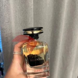 Bella Rouge parfym från RiiFFS - Snygg parfymflaska från RiiFFS, Bella Rouge. Flaskan är genomskinlig med svart och guldigt lock och dekorerad med ett rött band runt halsen. Inuti syns svarta pärlor som ger en cool touch. Perfekt för dig som gillar stilrena och unika detaljer. Likt baccarat rouge