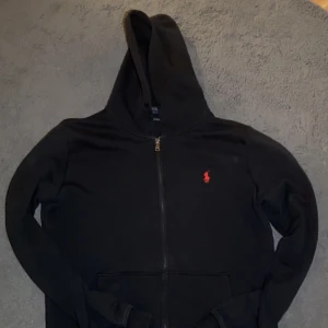 Svart hoodie från Polo Ralph Lauren - Svart hoodie från Polo Ralph Lauren med röd broderad logga på bröstet. Tröjan har huva, hel dragkedja och två fickor framtill. Tillverkad i mjuk bomull, perfekt för chill dagar. Klassisk och stilren design som funkar till det mesta.