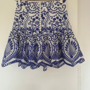 Vit och blå broderad kortkjol från Zara - Supersöt vit kortkjol med blått broderat mönster och dekorativa knappar framtill. Kjolen har volangkant och ett unikt, detaljerat blommönster som ger en cool och fräsch vibe. Perfekt för dig som gillar statement-plagg med mycket detaljer💕💕