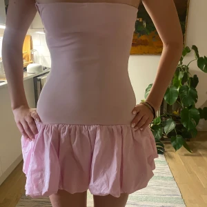 Ljusrosa axelbandslös kort klänning - Säljer en söt ljusrosa axelbandslös kort klänning med volang nedtill. Klänningen har en tight passform upptill och en fluffig kjol. Materialet känns som bomull och polyester, och den stängs med dragkedja i sidan. Dragkedjan var tyvärr sönder när jag köpte den, men är bara använd en gång och är utöver det i nyskick. 