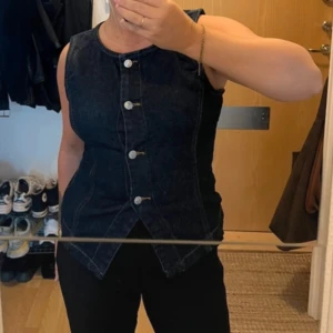 Mörkblå jeansväst med knappar - Säljer en snygg mörkblå jeansväst med markerade sömmar och silverfärgade knappar framtill. Västen har en figurnära passform och coolt snitt nertill som ger en edgy look. Perfekt att styla över en t-shirt eller på egen hand för en trendig vibe.