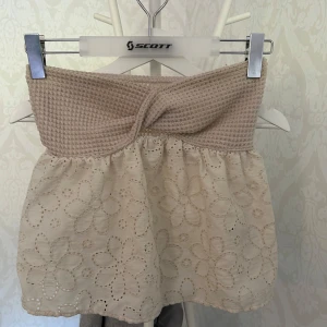 Beige tubtopp med virkad spets - Supersöt beige tubtopp med virkad överdel och broderad spets nertill. Toppen har ett twistat parti framtill och stora blommor i spetsen som ger en romantisk vibe. Perfekt till sommaren och lätt att matcha med jeans eller kjol.