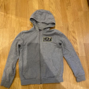 Grå EA7 hoodie med dragkedja - Snygg grå hoodie från EA7 Emporio Armani med gulddetaljer på bröstet. Tröjan har huva, hel dragkedja och två fickor framtill. Perfekt för dig som gillar stilrena och sportiga plagg med exklusiv känsla.