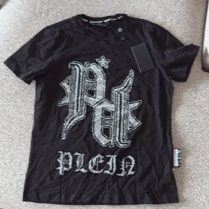 Svart Philipp Plein t-shirt med bling - Svart t-shirt från Philipp Plein med stort glittrigt logotryck framtill och coola detaljer. Klassisk rund halsringning och korta ärmar. Materialet är mjukt och skönt, perfekt för dig som gillar statement-plagg och streetstyle.
