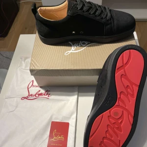Christian Louboutin svarta sneakers - Snygga svarta sneakers från Christian Louboutin i mocka med klassisk röd sula. Skorna har rund tå, svarta skosnören och diskreta metalldetaljer vid snörningen. Insidan är beige och mjukt fodrad för extra komfort. Perfekt för dig som vill sticka ut med exklusiv stil.