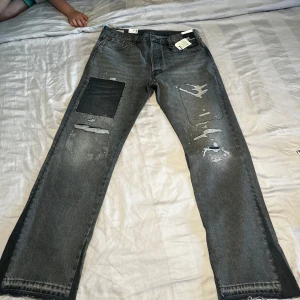 Grå Levis 501 jeans med patch och slitningar - Säljer ett par grå Levis 501 jeans med raka ben, coola slitningar och en svart patch på vänster lår. Jeansen har klassisk femficksdesign och är tillverkade i robust denim. Perfekta för dig som gillar en avslappnad och trendig streetstil. Storlek 33/32