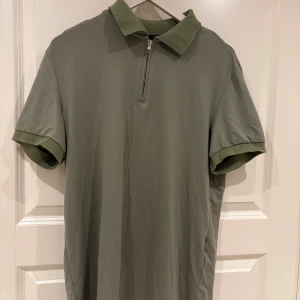 Olivgrön t-shirt med krage och zip - Snygg olivgrön t-shirt med krage och halv dragkedja framtill. T-shirten har korta ärmar med uppvikta muddar och är tillverkad i mjukt material. Perfekt för en chill och stilren look.