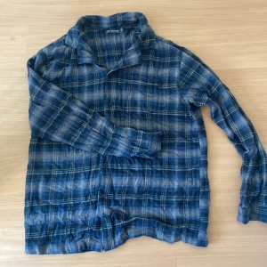 Blå och vit rutig skjorta - Säljer en blå och vit rutig skjorta med långa ärmar. Skjortan har en klassisk krage och är tillverkad i ett mjukt material. Perfekt för en avslappnad stil. (Pyjamas)