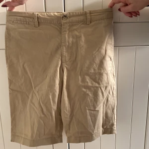 Beige shorts från Polo Ralph Lauren - Snygga beige shorts från Polo Ralph Lauren. De har en klassisk design med dragkedja och knapp framtill samt två bakfickor med knappar. Perfekta för en stilren och avslappnad look.