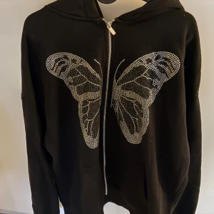 Svart hoodie med fjärilsdetalj från New Yorker - Snygg svart hoodie från New Yorker med en stor fjäril i glittrande stenar på framsidan. Den har en dragkedja och en bekväm huva. Perfekt för att ge din outfit lite extra bling! 🦋✨