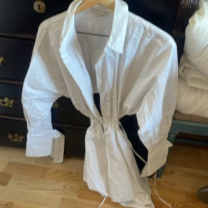 Vit kortklänning med knyt - Säljer en stilren vit kortklänning med knyt i midjan. Klänningen har långa ärmar och en klassisk krage. Perfekt för en chic och avslappnad look.