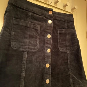 Mörkgrön manchesterkjol med knappar - Mörkgrön manchesterkjol med knappar framtill och praktiska fickor. Lite tecken på användning baktill i manchestermönstret