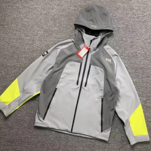 Helly Hansen - Säljer en grå och gul jacka från Helly Hansen med huva och dragkedja. Jackan har långa ärmar med neongula detaljer och flera praktiska fickor. Perfekt för utomhusaktiviteter.