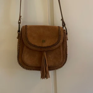 Brun axelväska i mocka med fransar - Brun  axelväska i mocka från Superdry med flätade detaljer och en dekorativ frans på framsidan. Väskan har ett lock med en liten metallknapp och justerbar axelrem. Perfekt för en bohemisk stil!        25x22cm
