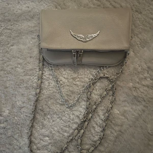Beige axelväska med kedja - Snygg beige axelväska från Zadig & Voltaire med en cool kedja som axelrem. Väskan har en dekorativ vingdetalj på framsidan och en praktisk dragkedja. Perfekt för att ge din outfit en stilfull touch.