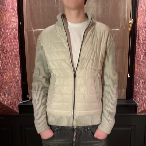 Beige och grå stickad cardigan med quiltad front - Snygg beige jacka med stickade ärmar och rygg, samt quiltad front. Jackan har en dragkedja framtill och ribbade muddar. Perfekt för kyligare dagar. 
