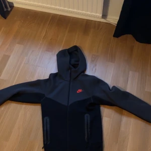 Svart hoodie från Nike - Säljer en svart hoodie från Nike med röd logga på bröstet. Den har en dragkedja framtill och två fickor med dragkedjor. Perfekt för en sportig look eller avslappnade dagar.