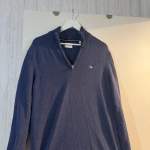 Blå tröja med dragkedja från GANT - Säljer en stilren blå tröja från GANT med dragkedja och broderad logotyp på bröstet. Tröjan har en ribbad krage och är perfekt för både vardag och avslappnade tillfällen. Passar utmärkt till jeans eller chinos.