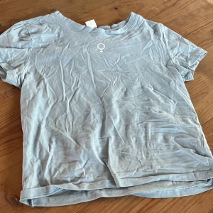 Ljusblå t-shirt med symbol - Säljer en ljusblå t-shirt med en liten symbol på bröstet. T-shirten har en rund hals och korta ärmar. 