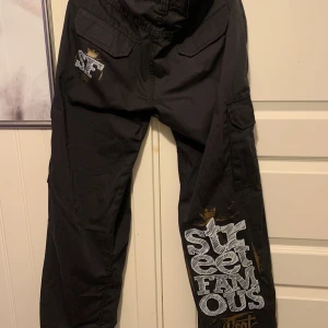 Svarta byxor från Bsat - Snygga svarta byxor från Bsat med trycket 'Street Famous' i vitt och guld på benet. Byxorna har flera fickor och en knappstängning. Perfekta för en avslappnad streetwear-look.