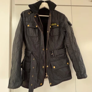 Svart jacka från Barbour - Säljer en snygg svart quiltad jacka från Barbour med guldiga knappar och dragkedja. Jackan har ett bälte i midjan och flera praktiska fickor. Perfekt för höst och vår!
