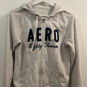 Grå hoodie från Aéropostale - Säljer en grå hoodie från Aéropostale med texten 'AERO Eighty Seven' i svart på framsidan. Tröjan har dragkedja och en bekväm huva med snören. Jättefint med ett par jeans 