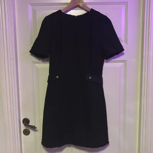 Svart klänning från H&M - Elegant svart klänning från H&M med korta ärmar och en stilren design. Klänningen har dekorativa knappar i midjan och en dragkedja i ryggen för enkel påklädning. Perfekt för en chic look.