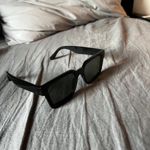 Svarta solglasögon med fyrkantig form - Snygga svarta solglasögon med en modern fyrkantig design. Perfekta för att ge en stilren look. De har breda skalmar och mörka linser som skyddar mot solen.