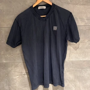 Svart t-shirt från Stone Island - Säljer en stilren mörkblå t-shirt från Stone Island med v-ringning och korta ärmar. T-shirten har en broderad logotyp på bröstet. Perfekt för en casual look.S fits M 8/10