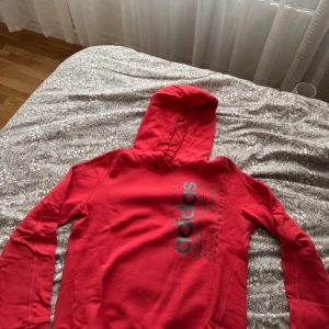 Röd hoodie från Adidas - Säljer en snygg röd hoodie från Adidas med svart texttryck på framsidan. Den har en klassisk huva och långa ärmar, perfekt för en avslappnad stil. Passar bra till både jeans och träningsbyxor.