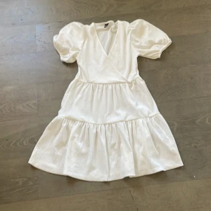 Vit omlottklänning med puffärmar - Säljer en söt vit omlottklänning med puffiga kortärmar och v-ringning. Klänningen har en luftig passform och volanger i kjolen, perfekt för sommarens varma dagar.Perfekt till studenten❤️