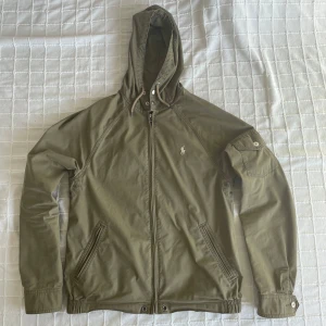 Olivgrön jacka från Ralph Lauren - Säljer en olivgrön/militärgrön jacka från Ralph Lauren med huva och dragkedja. Jackan har långa ärmar och broderad logotyp på bröstet. Perfekt för en stilren look. Den är limiterad om jag har rätt för mig men oavsett så är det en riktigt snygg jacka, passar perfekt som en overshirt eller som en windbreaker. Inga märkvärdiga skador, fläckar eller liknande, helt ärligt hade jag sagt att jackan är 9/10 sick. Sällan använt plagget, nypris 2700 kr. 