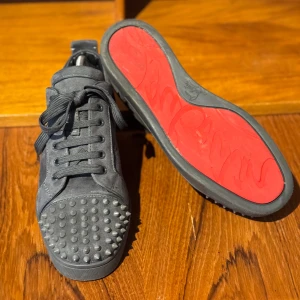Gråa Louboutin  - Snygga gråa sneakers med nitar på tån och röd sula. Skorna har snörning och är tillverkade i mocka. Perfekta för att ge din outfit en edgy touch. Storlek 42 men passar 43